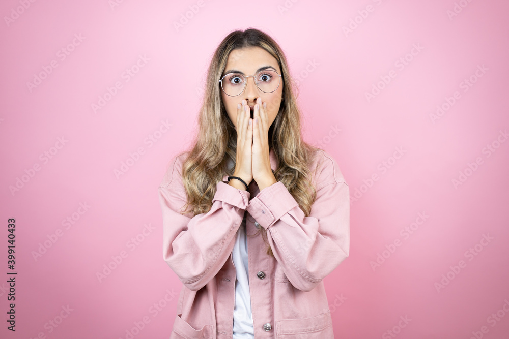 Young beautiful blonde woman with long hair standing over pink background with her hands over her mouth and surprised Young beautiful blonde woman with long hair standing over pink background with her hands over her mouth and surprised