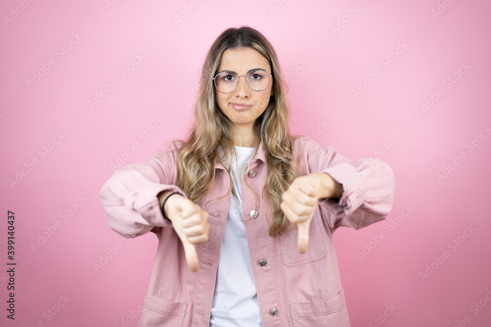 Young beautiful blonde woman with long hair standing over pink background with angry face, negative sign showing dislike with thumb down Young beautiful blonde woman with long hair standing over pink background with angry face, negative sign showing dislike with thumb down