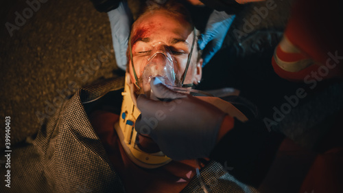 Portrait of a Young Injured Man Involved in a Traffic Accident is Being Saved by a Medical Team of EMS Paramedics on the Street at Night. Emergency Care Assistants Use a Ventilation Mask. © Gorodenkoff
