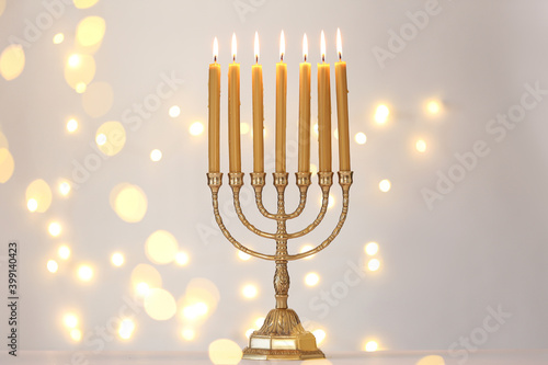 Golden menorah with burning candles against light grey background and blurred festive lights