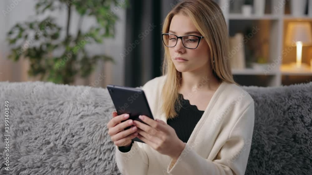 Attractive young woman in casual clothing and eyewear sitting on couch and surfing internet in digital tablet. Concept of people and modern technology. Attractive young woman in casual clothing and eyewear sitting on couch and surfing internet in digital tablet. Concept of people and modern technology.