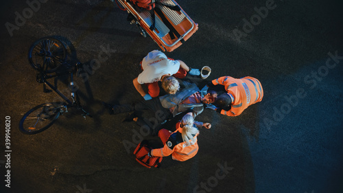 Aerial Portrait of a Young Injured Man Involved in Bicycle Accident is Being Saved by Medical Team of EMS Paramedics on the Street at Night. Emergency Care Assistants Provide Essential First Aid Help.
