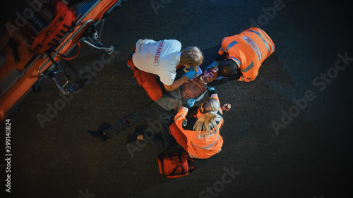 Canvas Print Aerial Portrait of a Young Injured Man Involved in Traffic Accident is Being Saved by Medical Team of EMS Paramedics on the Street at Night
