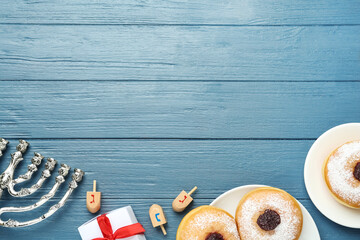 Flat lay composition with menorah, gift box, dreidels icluding He, Pe, Nun, Gimel letters and sufganiyot on blue wooden table, space for text. Hanukkah symbols