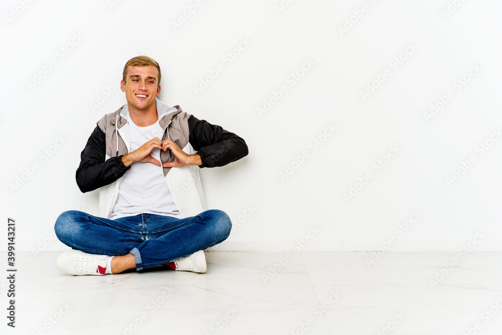 Young caucasian man sitting on the floor smiling and showing a heart shape with hands. Young caucasian man sitting on the floor smiling and showing a heart shape with hands.