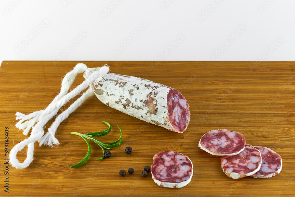 Spanish salami also sliced with black pepper and rosemary on a wooden board with white background. Copy space. Spanish salami also sliced with black pepper and rosemary on a wooden board with white background. Copy space.