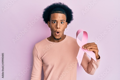 African american man with afro hair holding pink cancer ribbon scared and amazed with open mouth for surprise, disbelief face