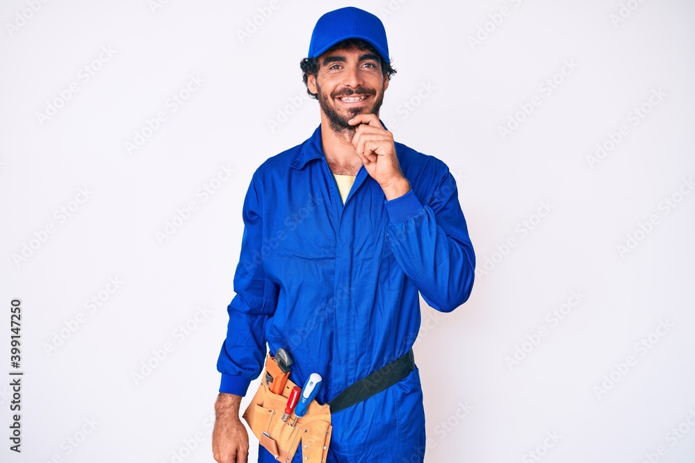 Handsome young man with curly hair and bear weaing handyman uniform looking confident at the camera smiling with crossed arms and hand raised on chin. thinking positive. Handsome young man with curly hair and bear weaing handyman uniform looking confident at the camera smiling with crossed arms and hand raised on chin. thinking positive.