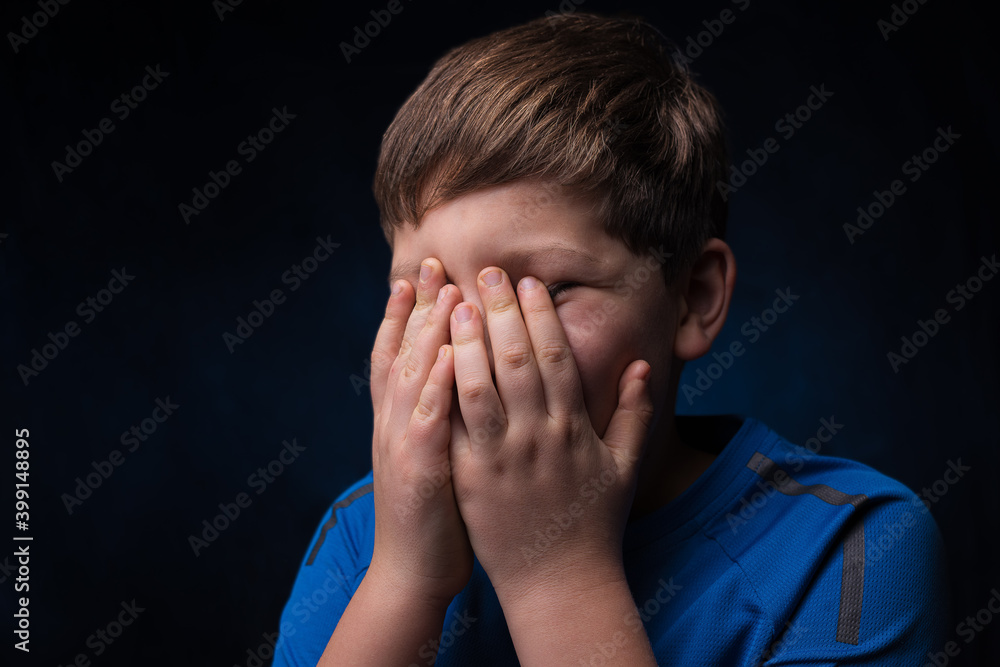white teenage boy with light brown hair in a blue sports t-shirt covering his face with two hands on  isolated background