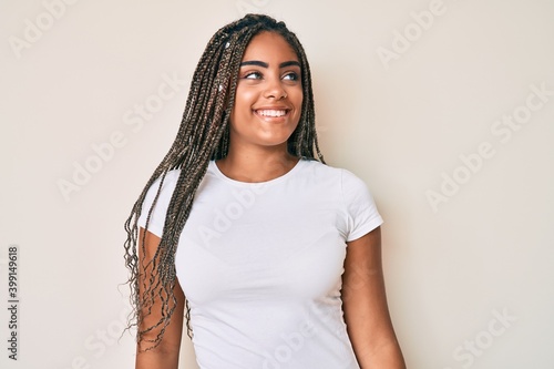 Young african american woman with braids wearing casual white tshirt looking away to side with smile on face, natural expression. laughing confident.