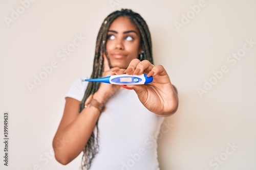 Young african american woman with braids holding thermometer serious face thinking about question with hand on chin, thoughtful about confusing idea