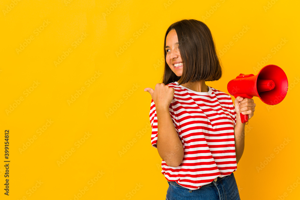 Young hispanic woman holding a megaphone points with thumb finger away, laughing and carefree. Young hispanic woman holding a megaphone points with thumb finger away, laughing and carefree.