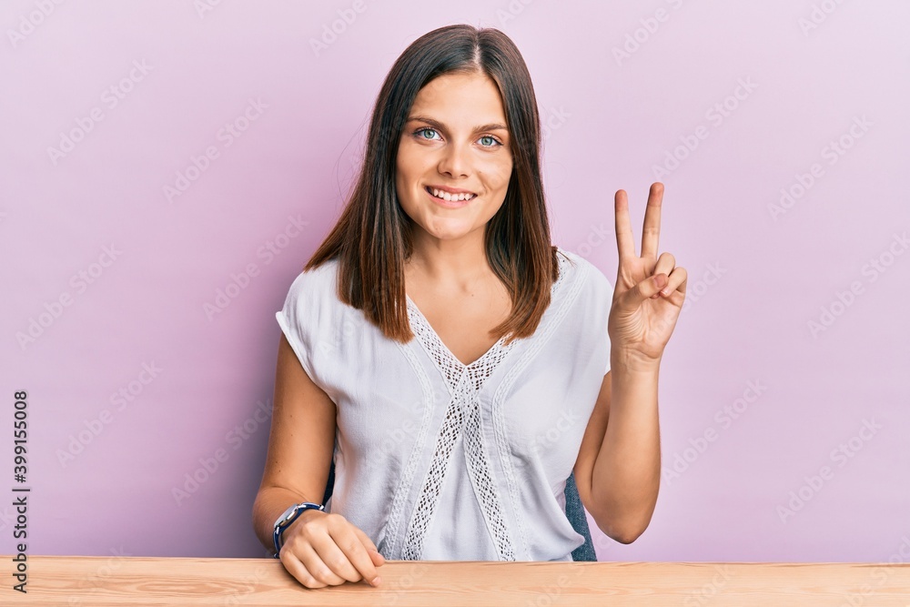 Young caucasian woman wearing casual clothes sitting on the table smiling with happy face winking at the camera doing victory sign. number two. Young caucasian woman wearing casual clothes sitting on the table smiling with happy face winking at the camera doing victory sign. number two.