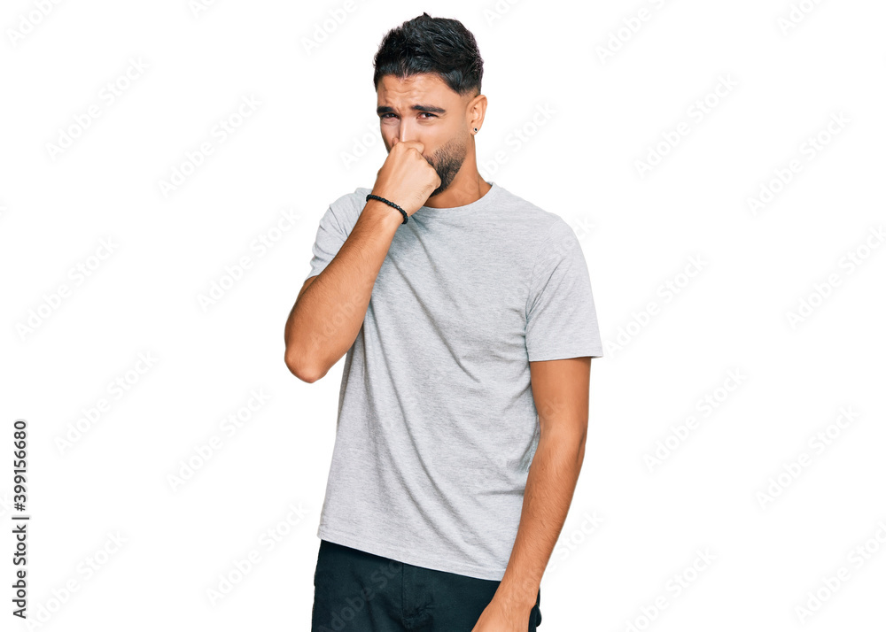 Young man with beard wearing casual grey tshirt smelling something stinky and disgusting, intolerable smell, holding breath with fingers on nose. bad smell Young man with beard wearing casual grey tshirt smelling something stinky and disgusting, intolerable smell, holding breath with fingers on nose. bad smell