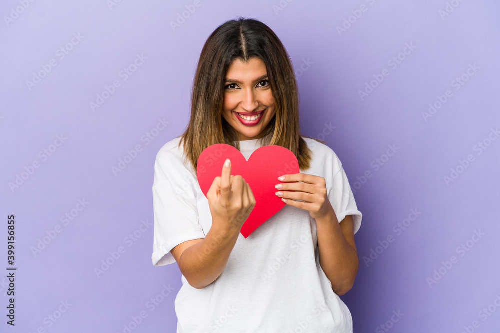 Young indian woman holding a valentines day heart isolated pointing with finger at you as if inviting come closer. Young indian woman holding a valentines day heart isolated pointing with finger at you as if inviting come closer.