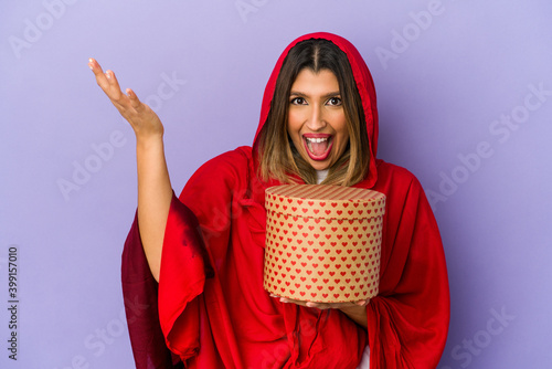 Young indian woman wearing a hijab holding a valentines day gift isolated receiving a pleasant surprise, excited and raising hands.