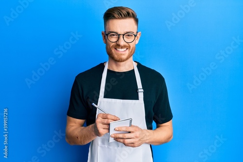 Young redhead man wearing waiter apron taking order smiling with a happy and cool smile on face. showing teeth. photo