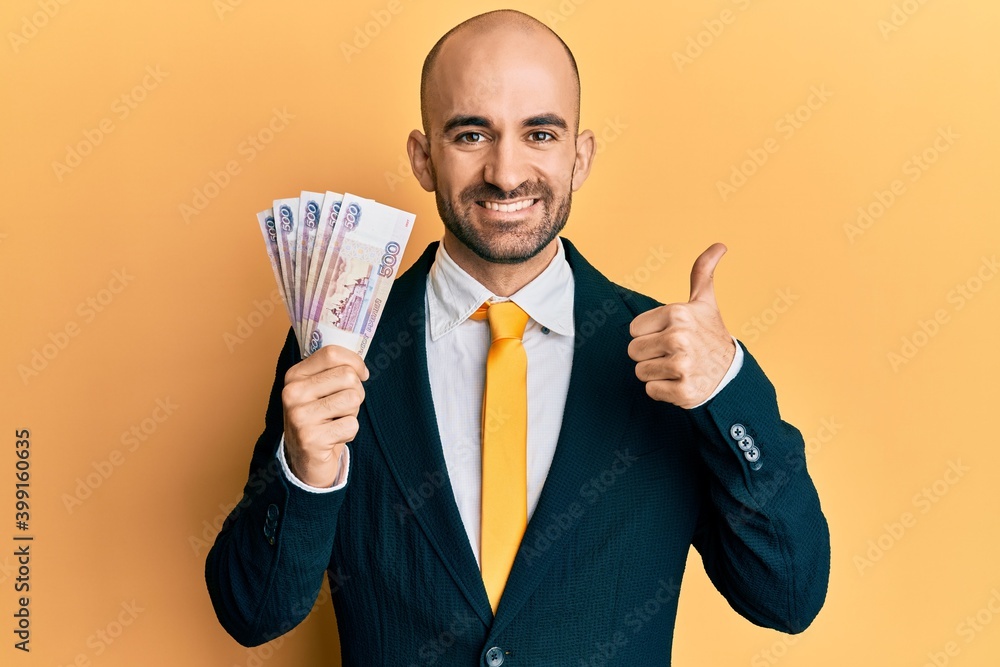 Young hispanic business man holding russian 500 ruble banknotes smiling happy and positive, thumb up doing excellent and approval sign Young hispanic business man holding russian 500 ruble banknotes smiling happy and positive, thumb up doing excellent and approval sign