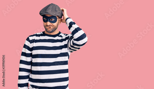 Young handsome man wearing burglar mask confuse and wondering about question. uncertain with doubt, thinking with hand on head. pensive concept.