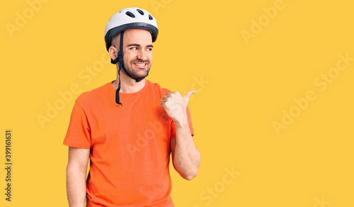 Young handsome man wearing bike helmet smiling with happy face looking and pointing to the side with thumb up.