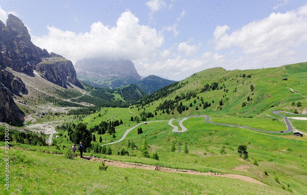 Hikers on a trail admire the view of a beautiful valley with a mountain highway winding thru green meadows ~ Summer scenery of majestic Dolomiti Mountains in Pass Gardena, Trentino, South Tyrol, Italy