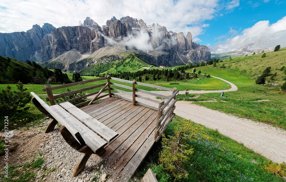An empty bench on a wooden deck with view of majestic Sella mountains & hiking tracks in the green grassy valley on a sunny summer day in Pass Gardena, Dolomiti, Bolzano, Trentino, South Tyrol, Italy