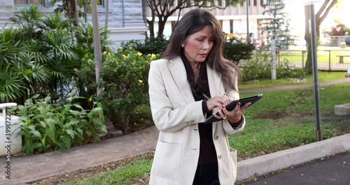 Attractive business woman with long hair and coat working on tablet with greenery background  Medium shot of successful older asian female working outdoors.