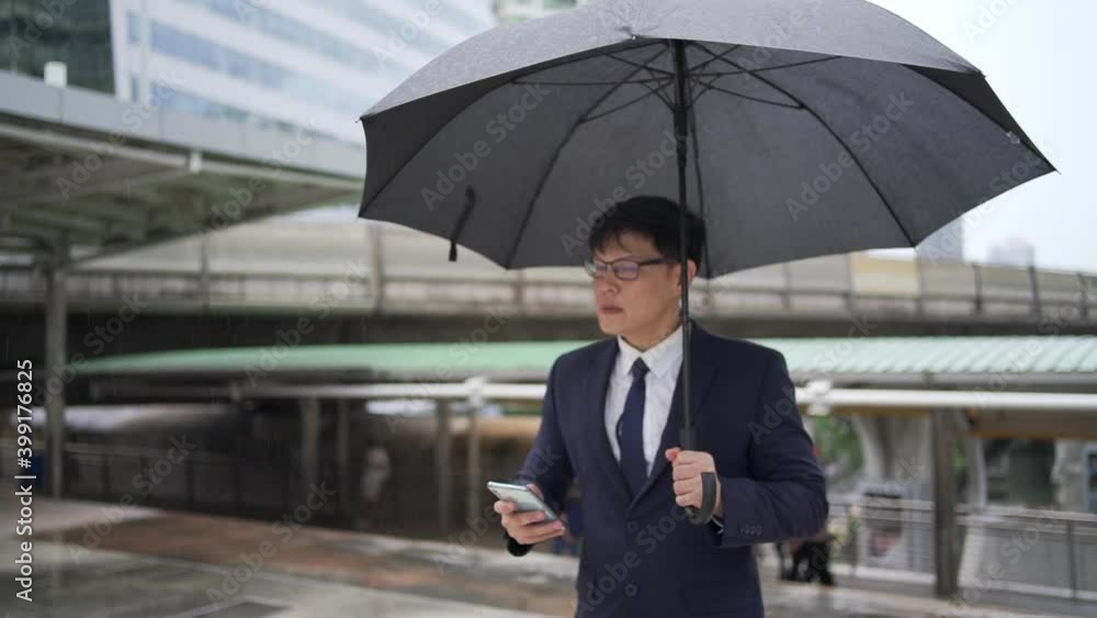 Confidence Asian businessman office worker in suit holding black umbrella walking in the rain with using smartphone. Handsome man business people working in city office district in bad weather day. Confidence Asian businessman office worker in suit holding black umbrella walking in the rain with using smartphone. Handsome man business people working in city office district in bad weather day.