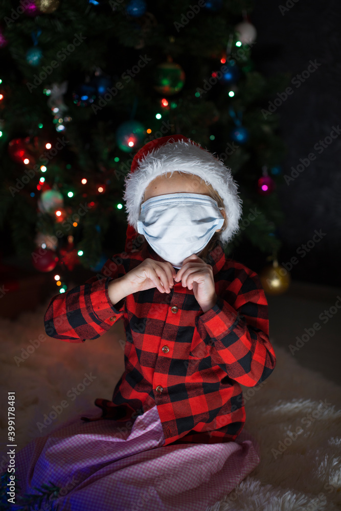 coronavirus, covid-19 in the new year. a little girl in a red Santa Hat and mask is sitting under the Christmas tree at home. coronavirus, covid-19 in the new year. a little girl in a red Santa Hat and mask is sitting under the Christmas tree at home.
