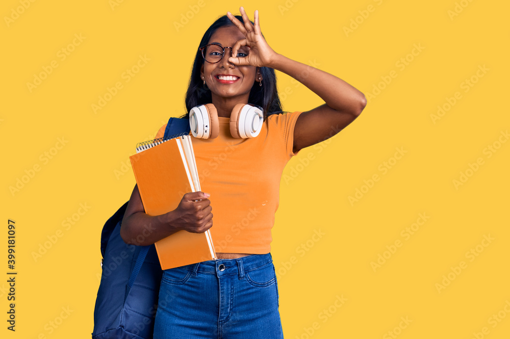 Young indian girl holding student backpack and books smiling happy doing ok sign with hand on eye looking through fingers Young indian girl holding student backpack and books smiling happy doing ok sign with hand on eye looking through fingers