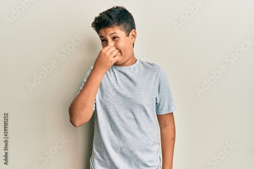 Teenager hispanic boy wearing casual grey t shirt smelling something stinky and disgusting, intolerable smell, holding breath with fingers on nose. bad smell