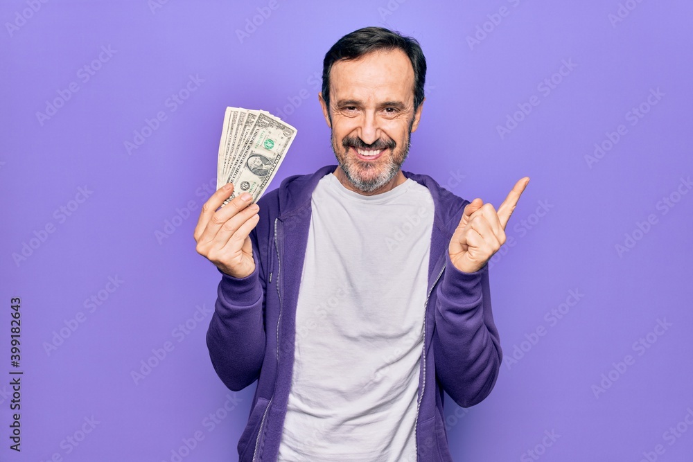 Middle age handsome man holding bunch of dollars banknotes over isolated purple background smiling happy pointing with hand and finger to the side Middle age handsome man holding bunch of dollars banknotes over isolated purple background smiling happy pointing with hand and finger to the side