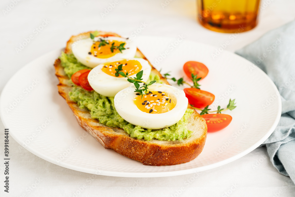 Avocado toast with toasted bread soft-boiled eggs with yellow yolk and tomatoes with herbs on white plate on light tablecloth