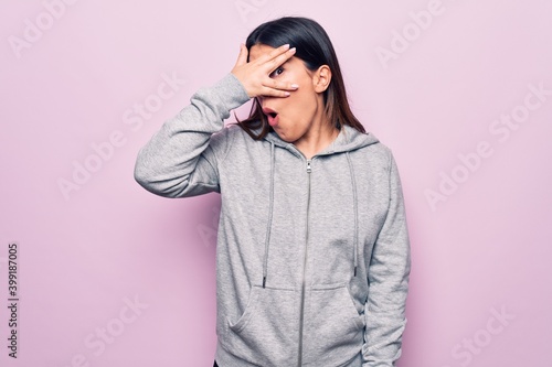 Young beautiful sporty woman wearing sportswear standing over isolated pink background peeking in shock covering face and eyes with hand, looking through fingers afraid © Krakenimages.com