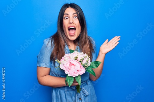 Young beautiful brunette woman holding bouquet of pink flowers over isolated blue background celebrating achievement with happy smile and winner expression with raised hand