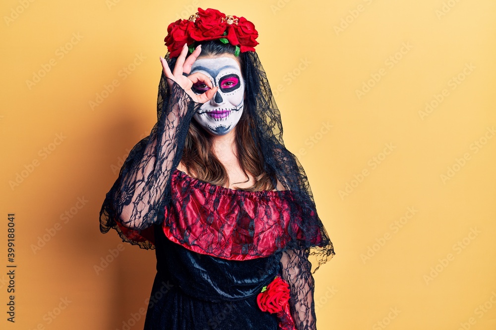 Young woman wearing day of the dead costume over yellow doing ok gesture with hand smiling, eye looking through fingers with happy face.