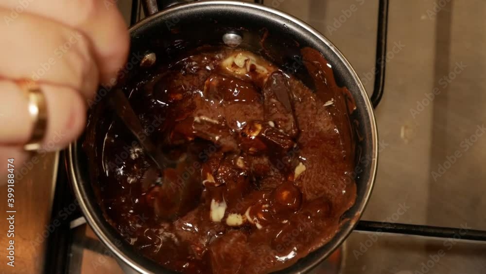 A broken milk chocolate bar with hazelnuts mixed with butter is being melted on the gas stove while a female with marriage ring is stirring in a small pot for melting. Above close up shot. A broken milk chocolate bar with hazelnuts mixed with butter is being melted on the gas stove while a female with marriage ring is stirring in a small pot for melting. Above close up shot.