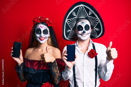 Couple wearing day of the dead costume holding smartphone showing screen smiling happy and positive, thumb up doing excellent and approval sign © Krakenimages.com