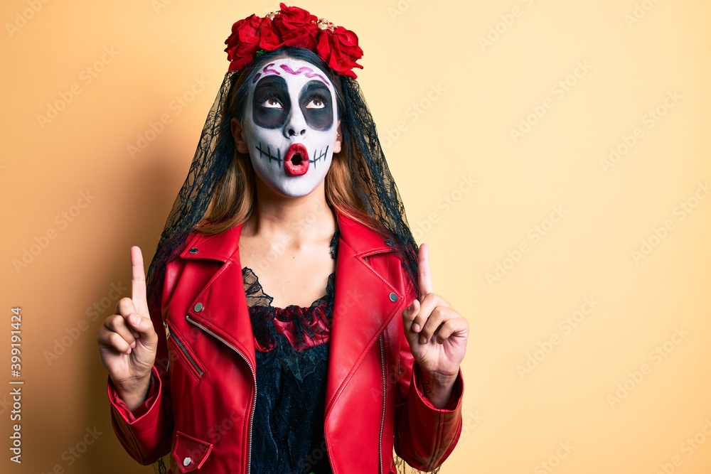 Woman wearing day of the dead costume over yellow amazed and surprised looking up and pointing with fingers and raised arms.