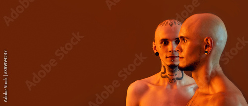 Front and side view of young half naked twin brothers with tattoos and piercings posing together, standing isolated over orange background