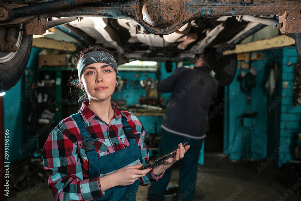 Portrait of a young female mechanic in uniform, posing with a tablet in her hands, standing under a car on a lift. Worker in the background blur