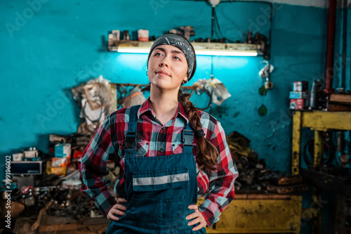 Portrait of a young beautiful female mechanic in uniform who poses with her hands on her hips and smiling. Working room in the background. The concept of gender equality