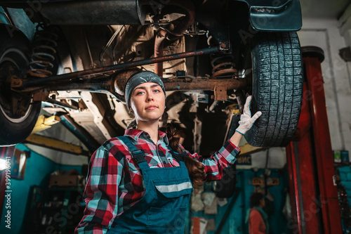 Portrait of a young female mechanic in uniform and gloves, posing near a car that is under repair. Bottom view. In the background is an auto repair shop