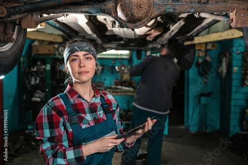 Portrait of a young female mechanic in uniform, posing with a tablet in her hands, standing under a car on a lift. Worker in the background blur