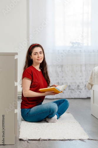 A young Caucasian smiling woman sits cross-legged on the floor and holds a book in her hands. In the background, the white interior of the room. The concept of education and reading books