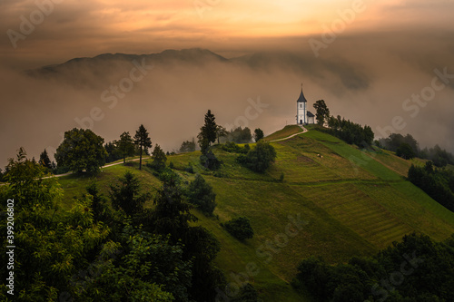 Jamnik, Slovenia - Magical foggy summer morning at Jamnik St.Primoz hilltop church. at sunrise The fog gently goes behind the small chapel with golden sky and Julian Alps at background