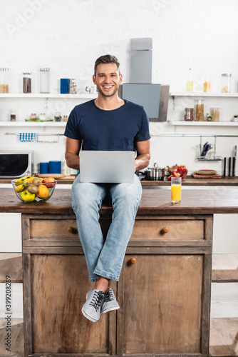 man looking at camera while using laptop near orange juice and fruits in kitchen