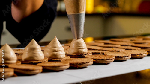 Chef is making caramel macarons. Hand with confectionery bag squeezing cream filling to macarons shells, close-up.
