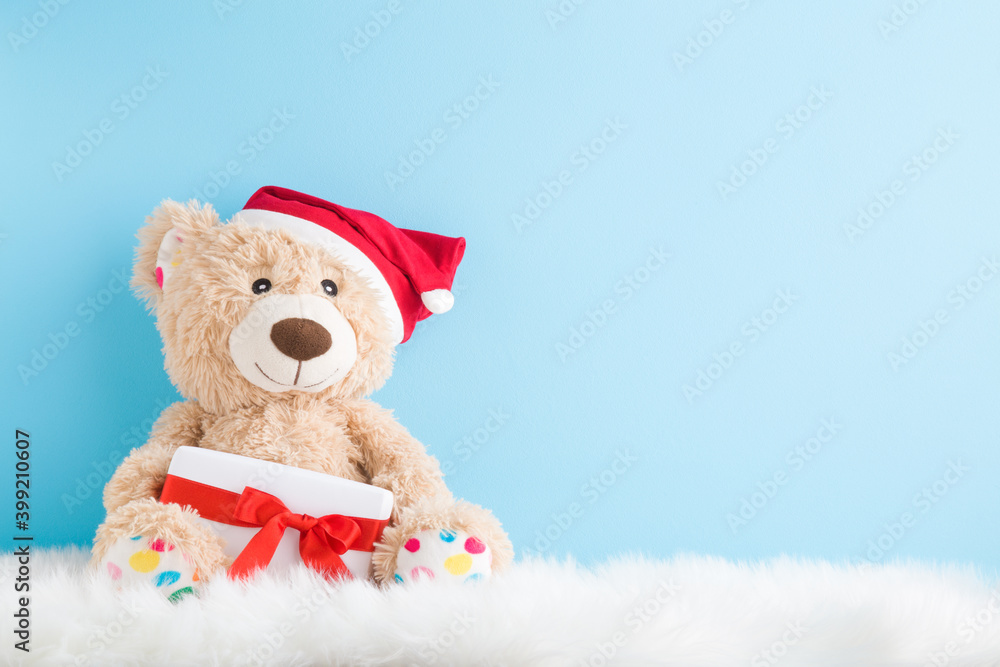 Smiling brown teddy bear with red hat sitting on fluffy fur blanket and holding white Christmas gift box with ribbon. Empty place for text, quote or sayings on light blue wall. Pastel color. Closeup.