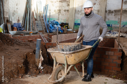 Young bearded man working at building site, carrying navvy wheelbarrow with bucket of construction mortar